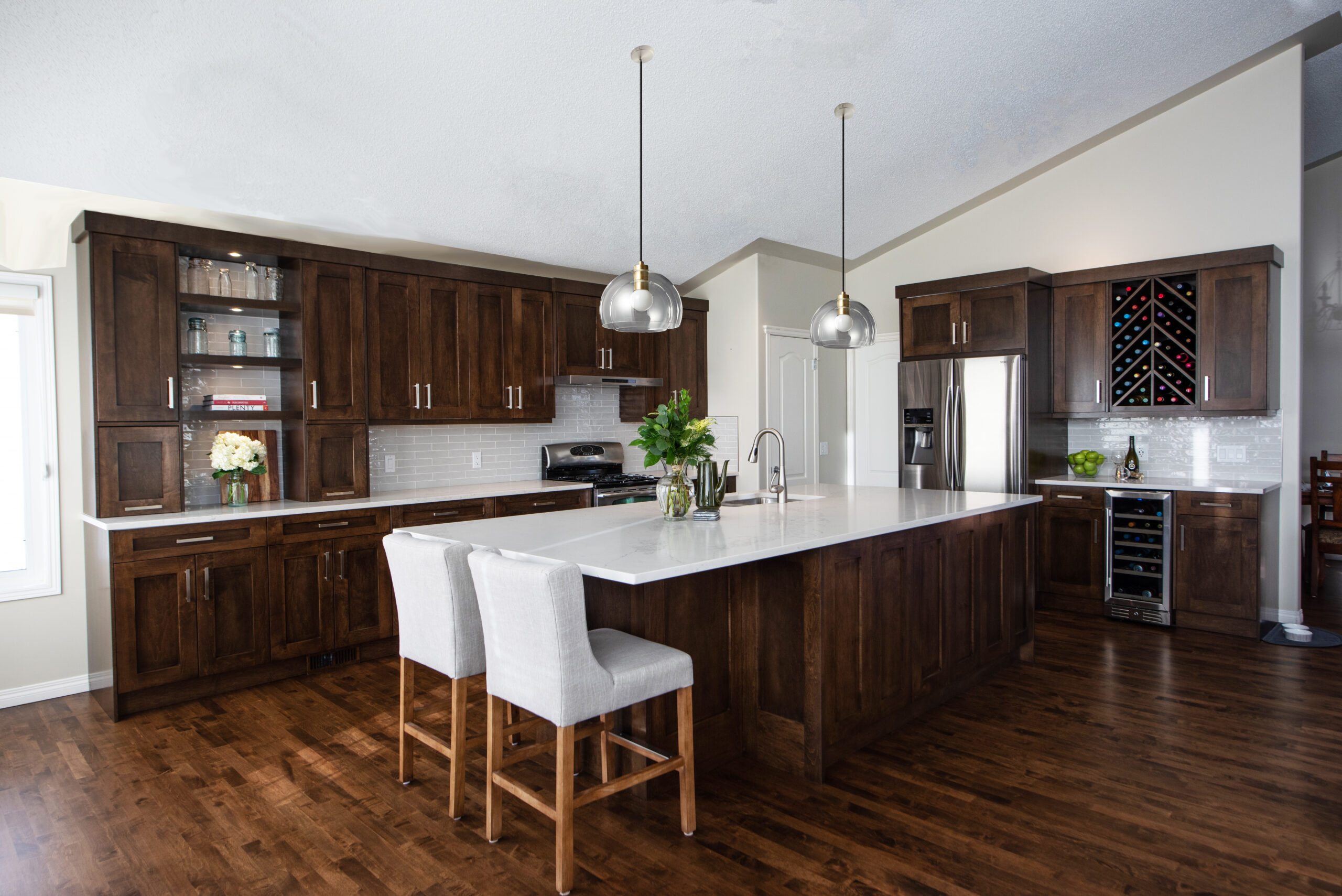 Custom kitchen renovation featuring rich dark wood cabinetry, a large quartz island with seating, stainless steel appliances, glass pendant lighting, built-in wine storage, and warm hardwood flooring.