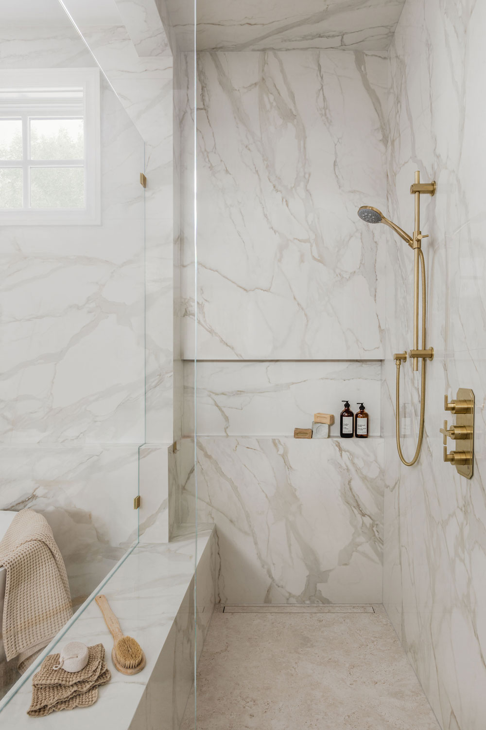 Luxury walk-in shower clad in large-format marble-look porcelain with frameless glass, built-in bench and niche, linear drain, and brushed brass handheld shower fixtures.