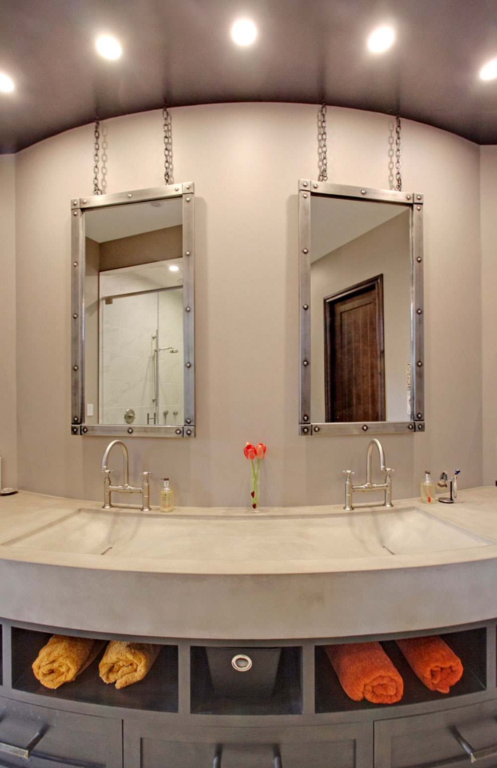 Before renovation: industrial-style double vanity with a curved concrete sink, dark cabinetry, open towel storage, metal-framed hanging mirrors and dark ceiling.