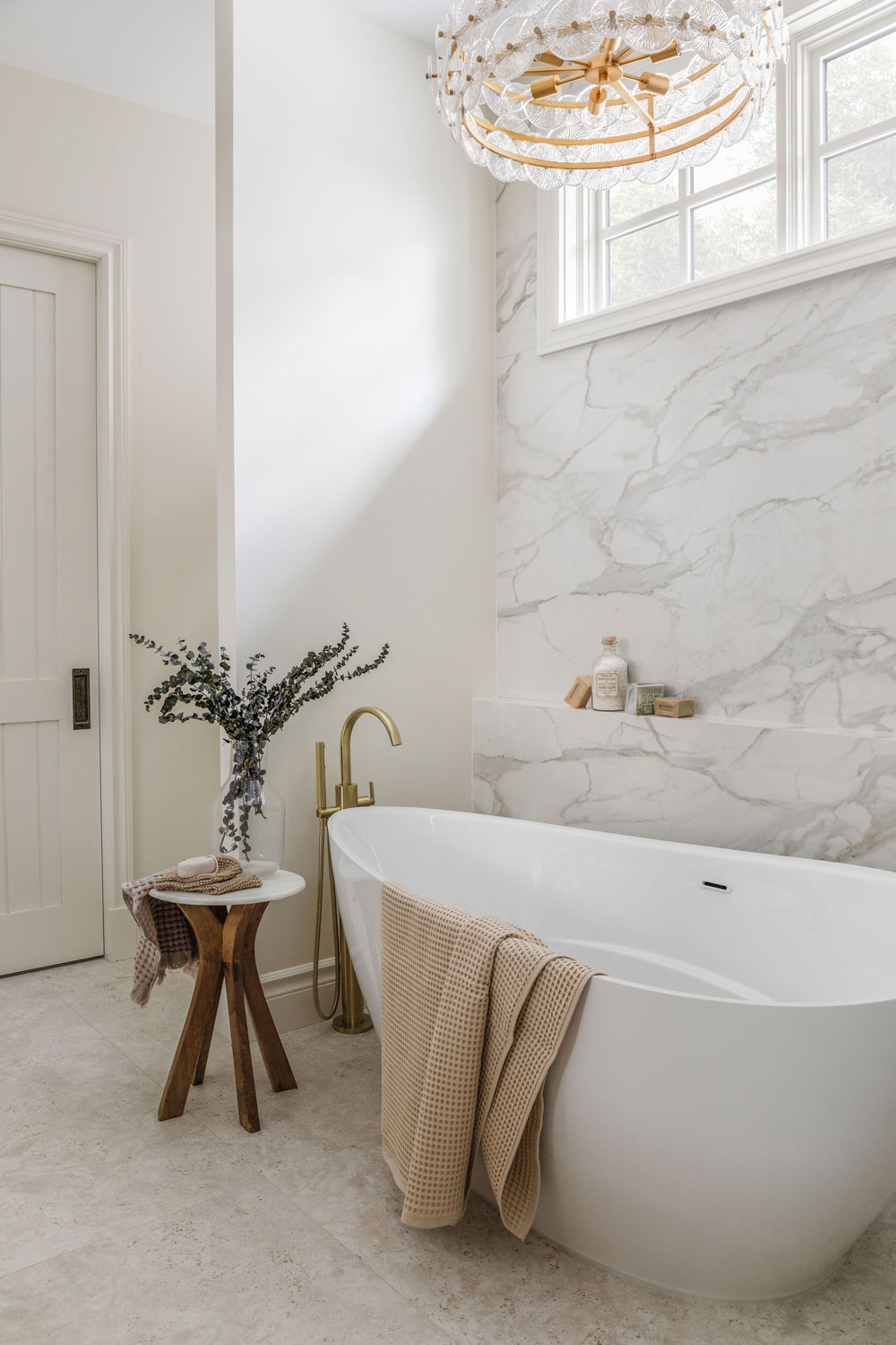 Spa-inspired ensuite with a freestanding soaker tub, brushed brass floor-mounted faucet, marble-look feature wall and statement glass chandelier.
