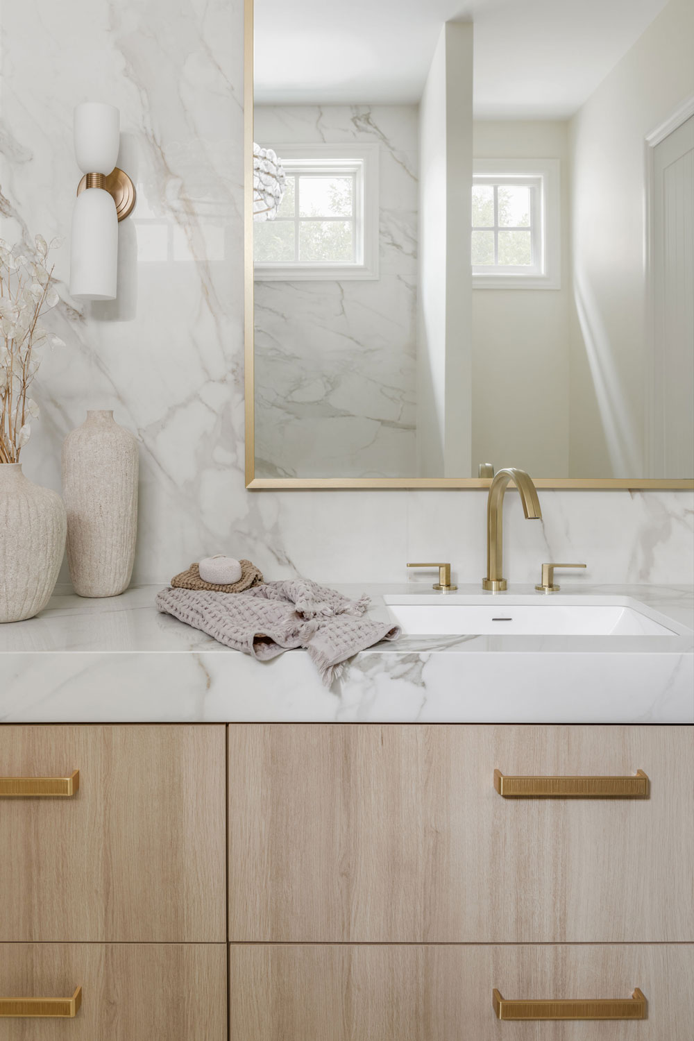 Close-up of a custom light wood ensuite vanity featuring a marble-look countertop and backsplash, brushed brass faucet and hardware, gold-framed mirror and modern wall sconce.