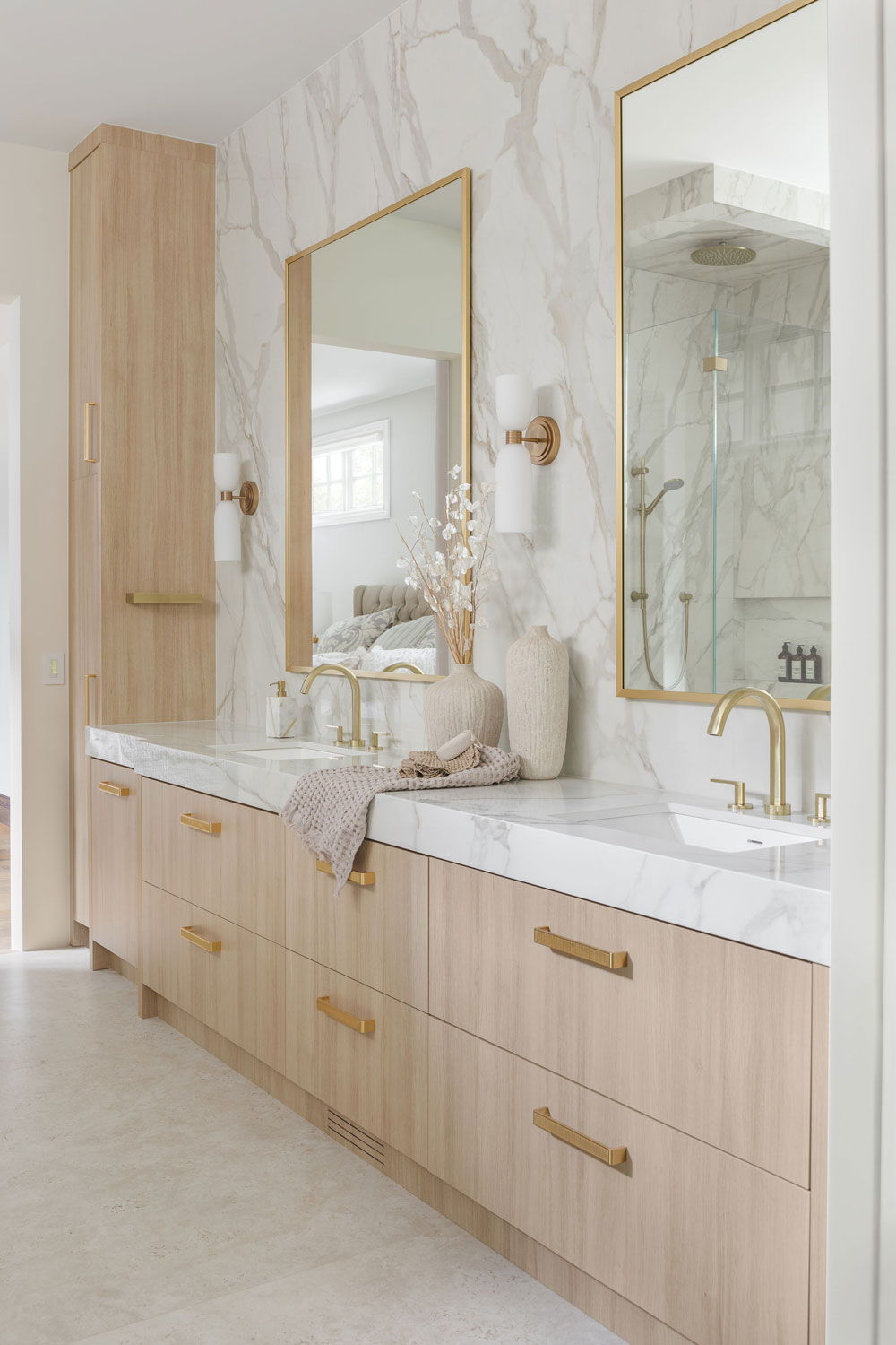Bright luxury ensuite featuring a custom light wood double vanity, marble-look porcelain feature wall, brushed brass fixtures, gold-framed mirrors and modern wall sconces.
