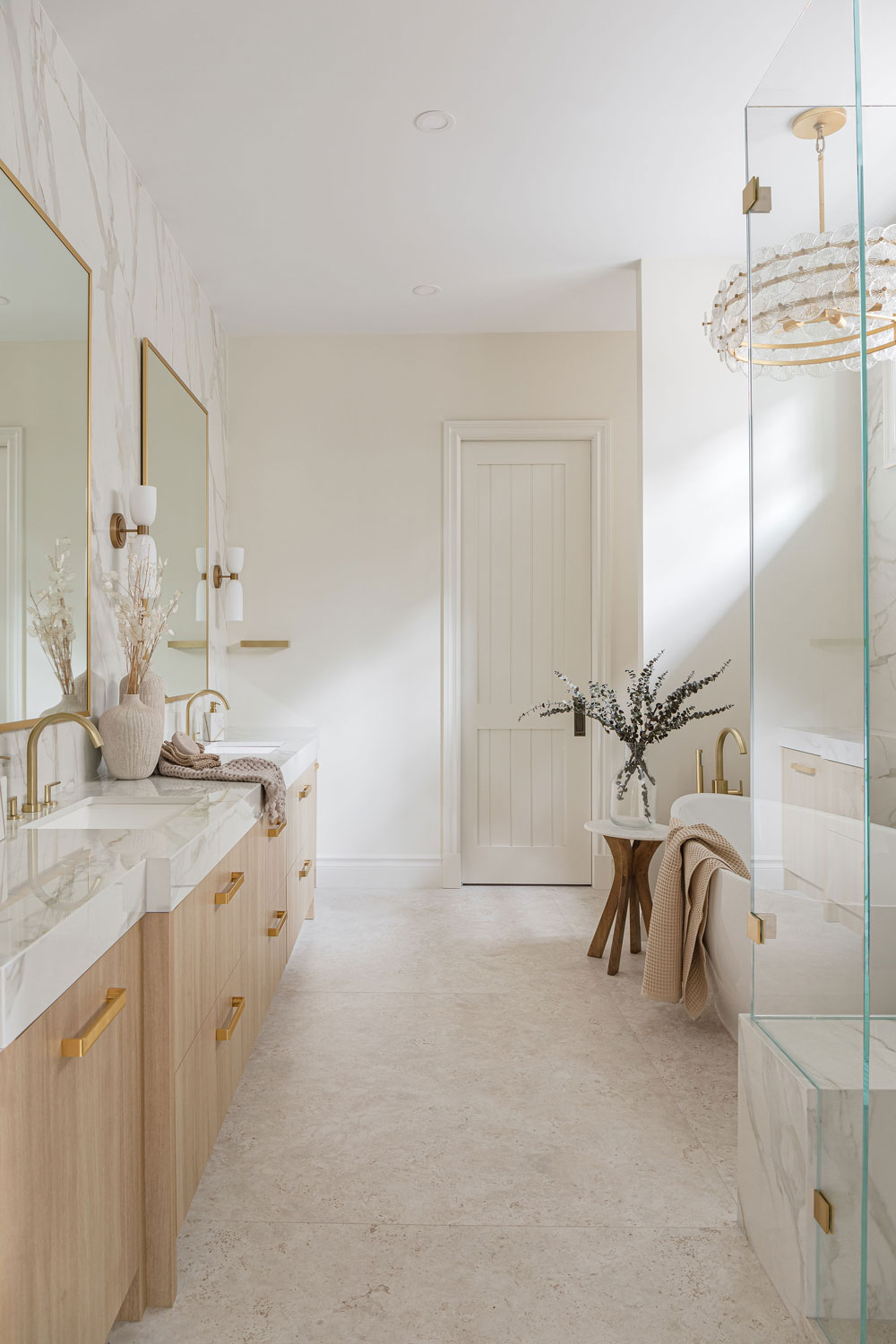 Bright spa-inspired ensuite renovation featuring a custom light wood double vanity, marble-look porcelain, brushed brass fixtures, freestanding tub and frameless glass shower.