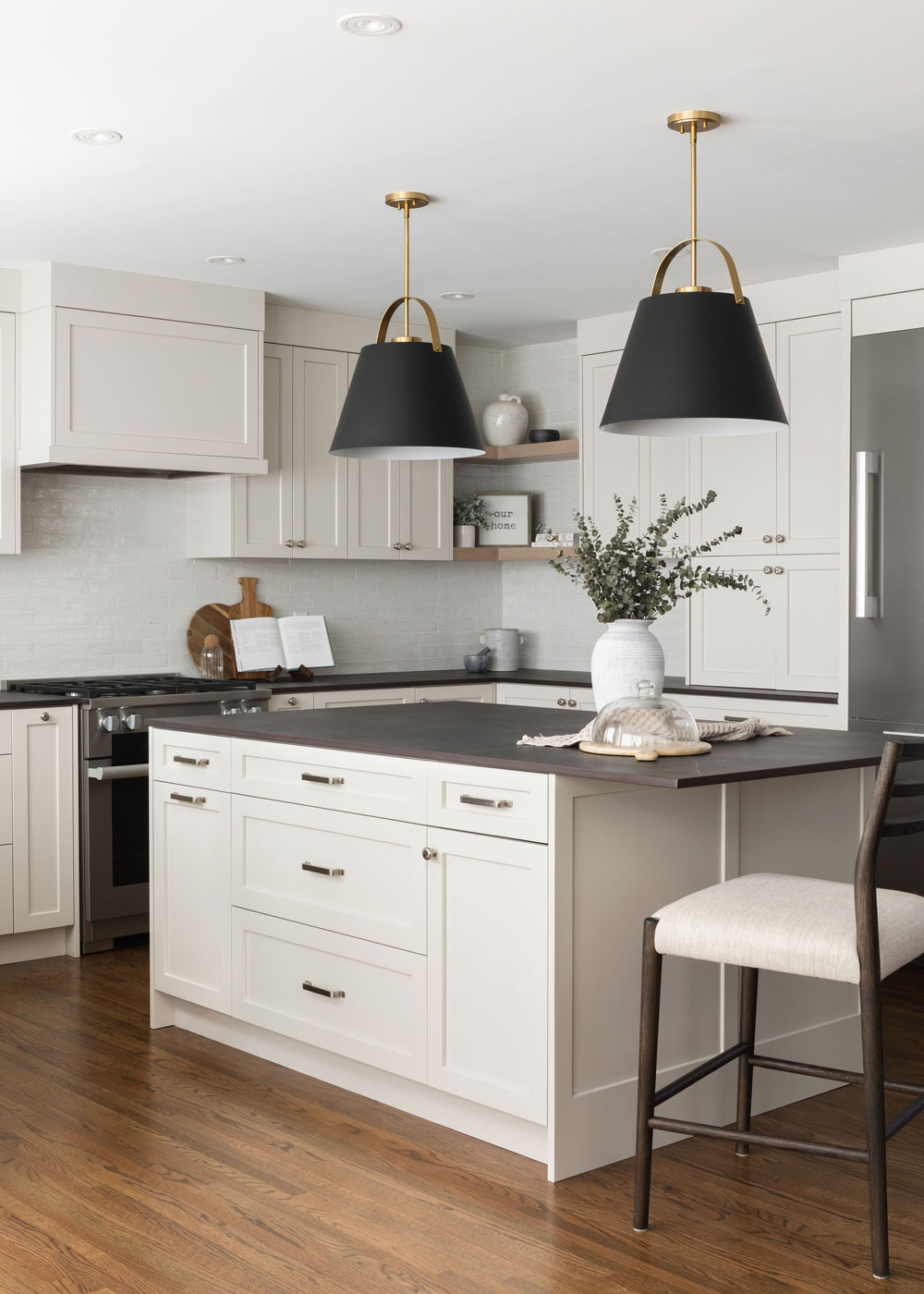 Modern kitchen with white cabinets, black pendant lights, and a central island featuring black and white bar stools. A vase with green foliage is on the counter.
