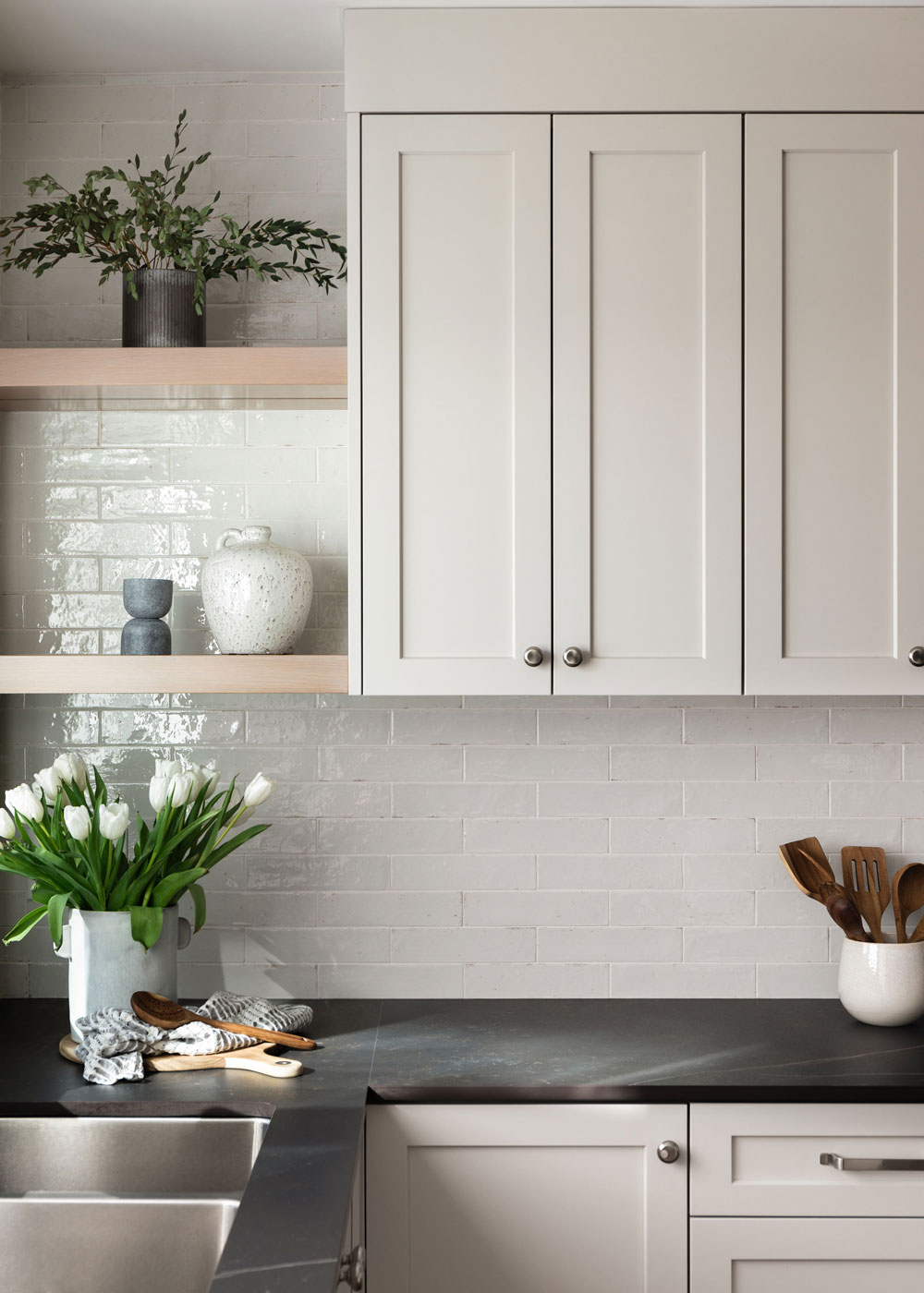 Modern kitchen with white cabinets, black pendant lights, and a central island featuring black and white bar stools. A vase with green foliage is on the counter.