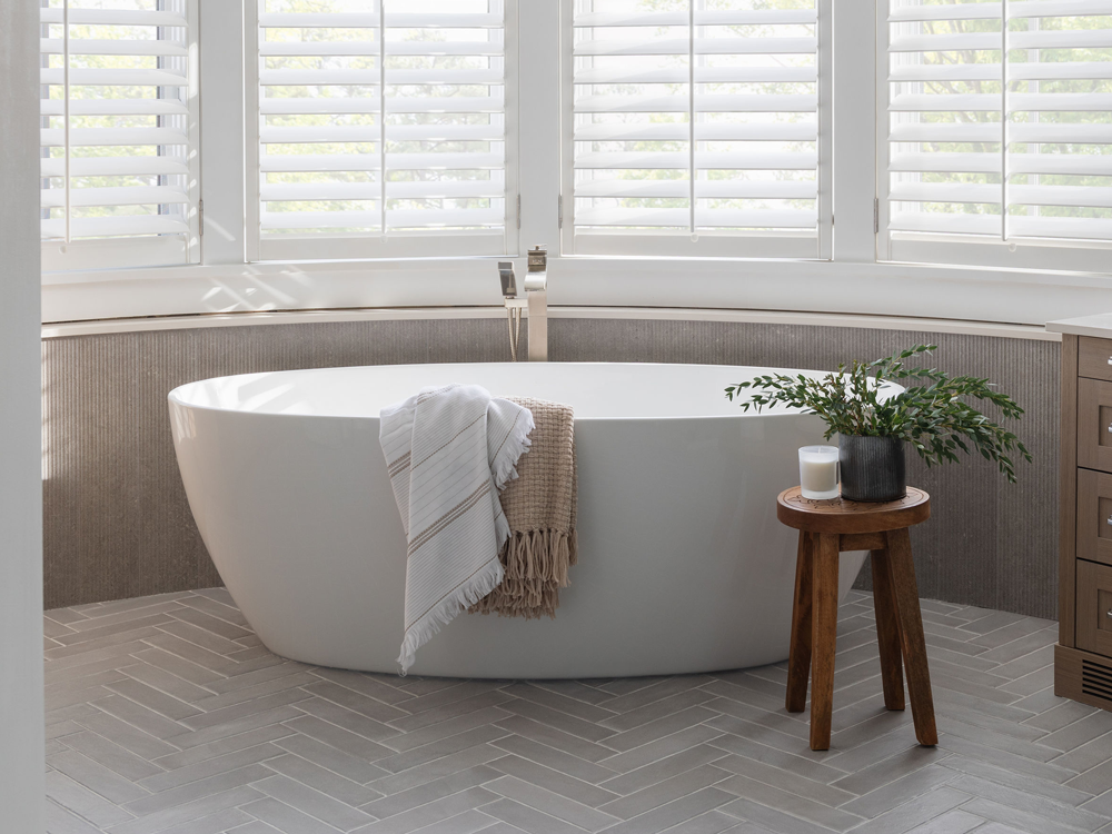 tub-cropped This bathroom exudes elegance and relaxation with its freestanding soaking tub placed in front of a curved wall of large windows, fitted with white plantation shutters that allow natural light to filter through. The combination of neutral tones, herringbone-patterned flooring, and textured wall paneling adds depth and sophistication. A wooden stool holding a potted green plant and a candle adds warmth, while draped towels over the tub enhance the inviting, spa-like atmosphere. The sleek floor-mounted faucet completes the modern yet timeless aesthetic.